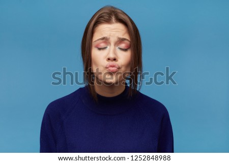 Close up portrait of young playful female, with dark hair and dressed casualy, keeps her eyes closed and kiss. Isolated over blue background