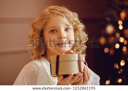 Charming elegant European baby with blonde hair and big blue eyes holds gift in small round box, on blur background glare from Christmas tree lights.