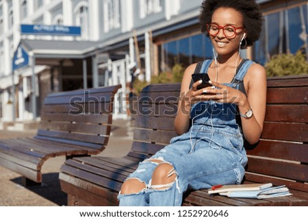 Photo of cheerful black woman holds cellular, types text messages, uses earphones, listens music, dressed in ragged overalls, models outdoor, enjoys playlist. Modern technologies, online communication