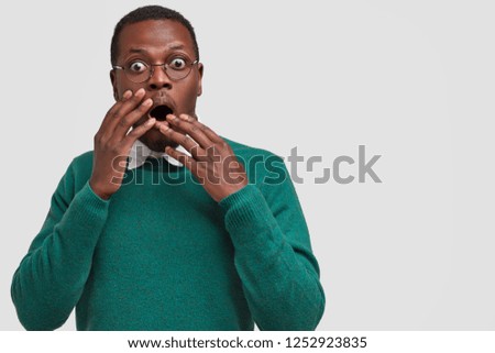 Photo of dismayed frightened black hipster covers mouth with both hands, wears green outfit, keeps eyes popped out, models over white background with copy space for your avertisement or text