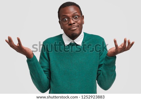 Hesitant uncertain man with dark skin, shrugs shoulders in bewilderment, has doubtful look, spreads palms, dressed in green sweater, isolated over white background. People and hesitation concept Royalty-Free Stock Photo #1252923883