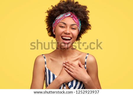 Positive dark skinned young woman with pleased expression, feels satisfied to hear compliment or confession in love, keeps both hands on chest, poses over yellow background, wears casual clothes Royalty-Free Stock Photo #1252923937