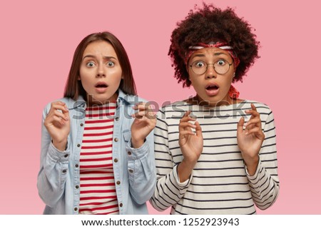 Photo of scared multiethnic women look with terror, notice something horrible, keep hands in protective gesture, wear striped clothes, isolated over pink background. People and fear concept.