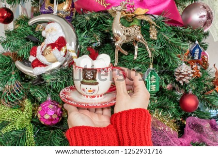 Women's hands in a wool sweater holding a mug with a snowman and marshmallow on the background of the Christmas tree. The concept of the Christmas holidays and the new year.