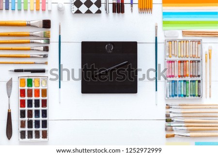 Supplies and devices for creative art work concept, supplies set and digital wacom tablet on white wooden background, paintbrushes paints crayons pencils on table, back to school. Top view copy space