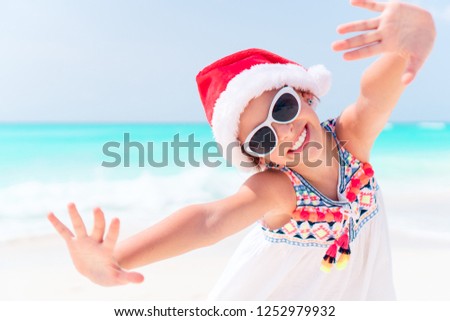 Little happy girl in Christmas hat on white beach having fun and looking at camera