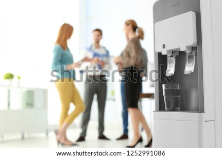 Modern water cooler with glass and blurred office employees on background. Space for text Royalty-Free Stock Photo #1252991086