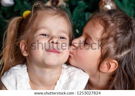 Two cute sisters of the same age lie next to the New Year tree. One kisses the other on the cheek. Concept of love and friendship of sisters and brothers, strong family and family relationships
