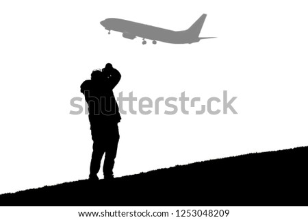 A black and white image of a silhouetted photographer on a hill taking a photo of a passenger airline that has taken off from an airport.