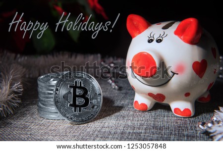 Winter season greeting card with few silver colored bitcoin coins, red and white pig money bank, Christmas decor and Happy holidays text. Selective focus. Investment and savings concepts.