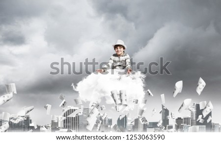 Young little boy keeping eyes closed and looking concentrated while meditating on cloud among flying papers with cityscape view on background.