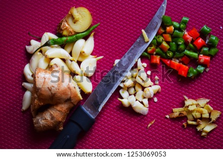 Stock photo of peeled  whole and chopped garlic, few green chillies and ginger  kept on purple color chopping board with knife. Picture captured under natural light at Bangalore, Karnataka, India.
