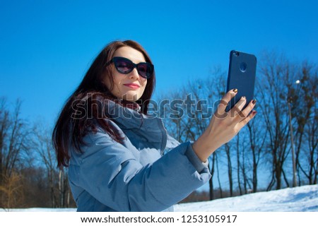 Beautiful brunette with long hair in sunglasses makes selfie against the blue sky and winter park. Waiting for spring, winter vacation