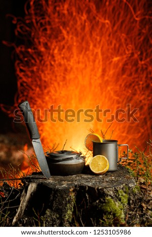 A mug, a teapot, a cut lemon and a knife stand on a stump in the forest at night. A bonfire with sparks is blurred in the background. Vertical frame