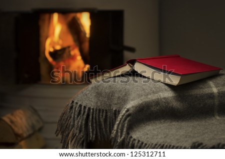 Peaceful image of open book resting on a arm rest of a couch. Warm fireplace on background.