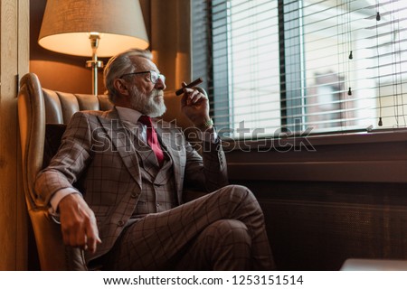 Serious old-aged businessman wearing classy custom tailored suit, red tie and expensive wristwatch sitting on old fashioned sofa in office, looking at camera and smoking cigar Royalty-Free Stock Photo #1253151514