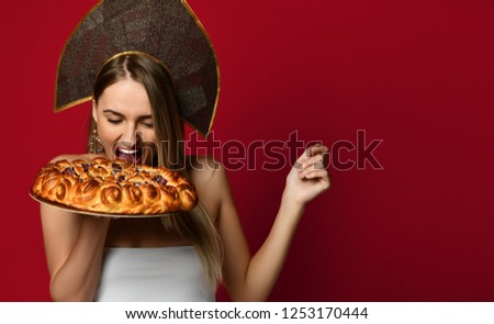 Portrait of young blonde woman in Russian traditional cap hat kokoshnik happy smiling bite eat sweet cherry homemade pie tasty bun looking up on red background