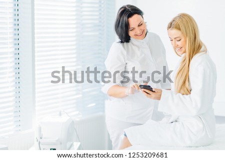 Beautiful blonde female in white bathrobe making appointment at beautician, checking vacant time at her shedule in smartphone