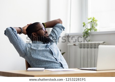 Black african millennial businessman take break during workday relaxing sitting on chair at office desk hold hands behind head looking at window thinking feels good. Stress relief daydreaming concept Royalty-Free Stock Photo #1253228191