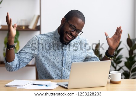 Joyful black businessman sitting at desk looking at computer screen talking with friend make informal video call. African worker received great news read email feels happy getting reward or promotion Royalty-Free Stock Photo #1253228203