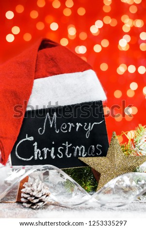 A table with the text "Merry Christmas" written in a chalk covered with Santa's hat on a wooden board covered with snow with a branch of fir, pinecone, decorative ribbon and a silver star.