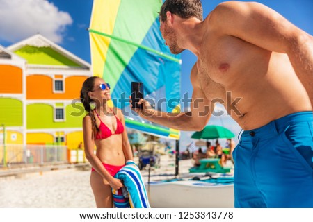 Beach vacation couple taking phone pictures - man taking pictures of model woman posing in bikini sun bathing in summer. Florida travel.