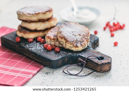 On a white table made of artificial stone is a wooden cutting board with five cottage cheese pancakes and red berries . On the table a red checkered napkin, sour cream, fork and knife.