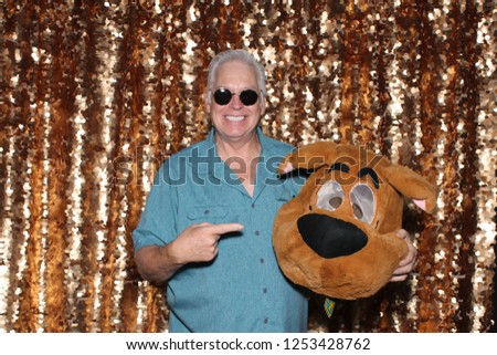 Photo Booth. A man poses and smiles with an cartoon animal head in a Photo Booth. Gold Sequin background. 