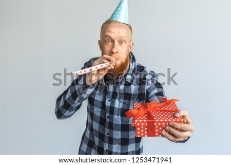 Red hair mature man wearing birthday cap standing isolated on grey wall giving gift box to camera close-up blowing party horn smiling cheerful