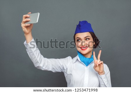 Young woman professional stewardess standing isolated on grey wall taking selfie photo on smartphone showing peace gesture looking camera smiling playful