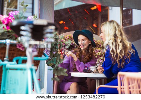Two smiling elegant young women sitting in the restaurant outdoor and looking at the phone