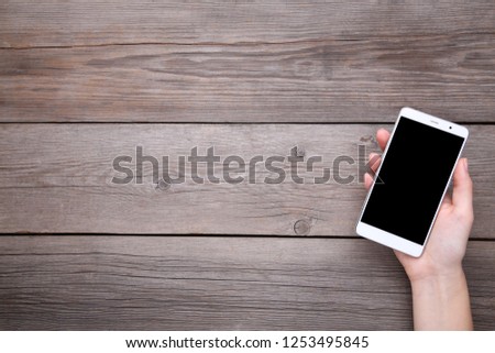 Female hands holding mobile phone with blank screen on grey wooden table. Smartfone white