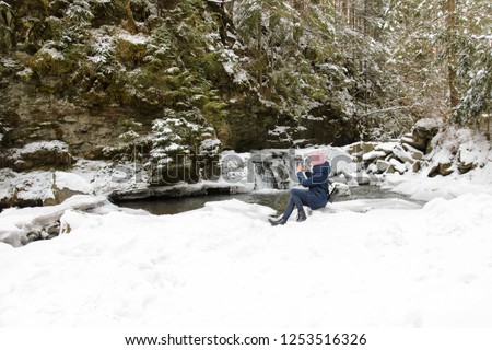Girl in blue coat sits and makes the photo on a smartphone on a background of icy waterfall and rocks in a snow-covered coniferous forest. Winter cloudy day.