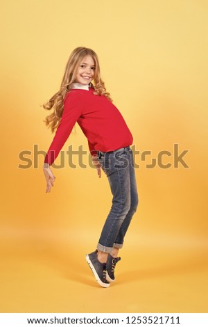 Kid fashion and style. Girl in red sweater and jeans stand on tiptoe. Beauty, look, hairstyle. Child smile with curly blond hair on orange background. Happy childhood concept.