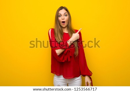 Young girl with red dress over yellow wall surprised and pointing side