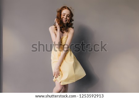 Slim pretty girl with red curly hair posing in studio with smile. Indoor shot of wonderful caucasian woman wears summer yellow dress.
