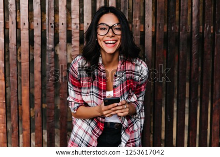Excited elegant girl in spring outfit laughing on wooden background. Outdoor shot of amazing latin woman in glasses holding phone with smile.