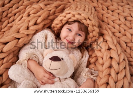 child in a hat under a blanket of natural sheep wool. Merino plaid knit covers little girl. designer hats natural wool yarn. crocheted dress. happy baby in a crib hugging a Teddy bear