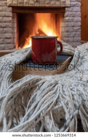 Red mug with tea, and warm wool scarf , a book , near cozy fireplace, in country house, winter vacation.