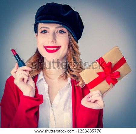 Portrait of young redhead woman in red cardigan and hat with lipstick and gift box on grey background. Image made with native lights
