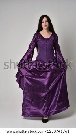 
full length portrait of beautiful girl with long black hair,   wearing purple fantasy medieval gown. standing pose on grey studio background.
