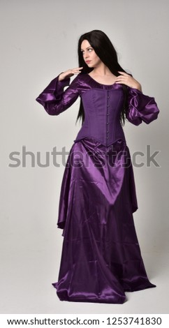 
full length portrait of beautiful girl with long black hair,   wearing purple fantasy medieval gown. standing pose on grey studio background.
