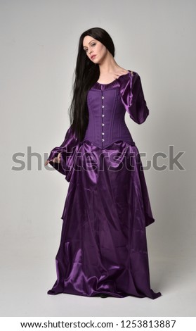 
full length portrait of beautiful girl with long black hair,   wearing purple fantasy medieval gown. standing pose on grey studio background.