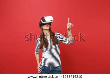 Young girl in virtual reality glasses touch something like push on button pointing at floating virtual screen isolated on red background. People sincere emotions lifestyle concept. Mock up copy space