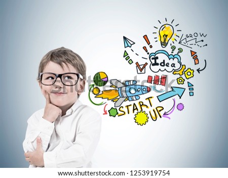 Smiling thinking little boy wearing glasses and white shirt standing near gray wall with colorful start up sketch drawn on it. Concept of future of business