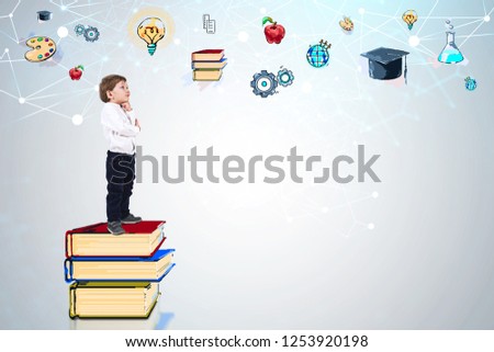 Side view of adorable little boy in white shirt and jeans thinking standing on stack of books near white wall with colorful education sketch. Mock up