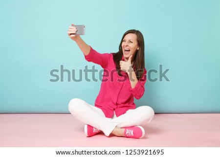 Full length woman in rose shirt, white pants sitting on floor doing selfie shot on cellphone isolated on bright pink blue pastel wall background studio. Fashion lifestyle concept. Mock up copy space