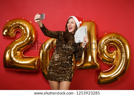 Merry Santa girl in shiny glitter dress Christmas hat hold money cellphone isolated on bright red background golden numbers air balloons studio portrait. Happy New Year 2019 holiday party concept