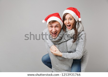 Merry fun couple girl guy in red Santa Christmas hat gray sweaters scarves isolated on grey wall background, studio portrait. Happy New Year 2019 celebration holiday party concept. Mock up copy space