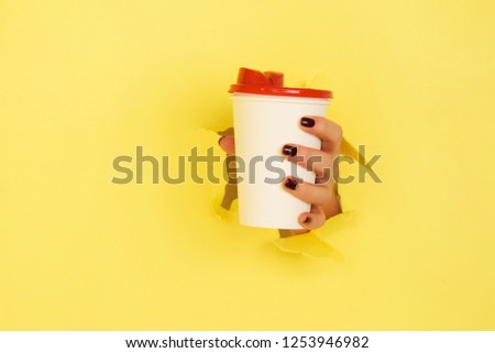 Female hand holding white paper mug on yellow background. Take away coffee cup concept. Mock up with copy space.